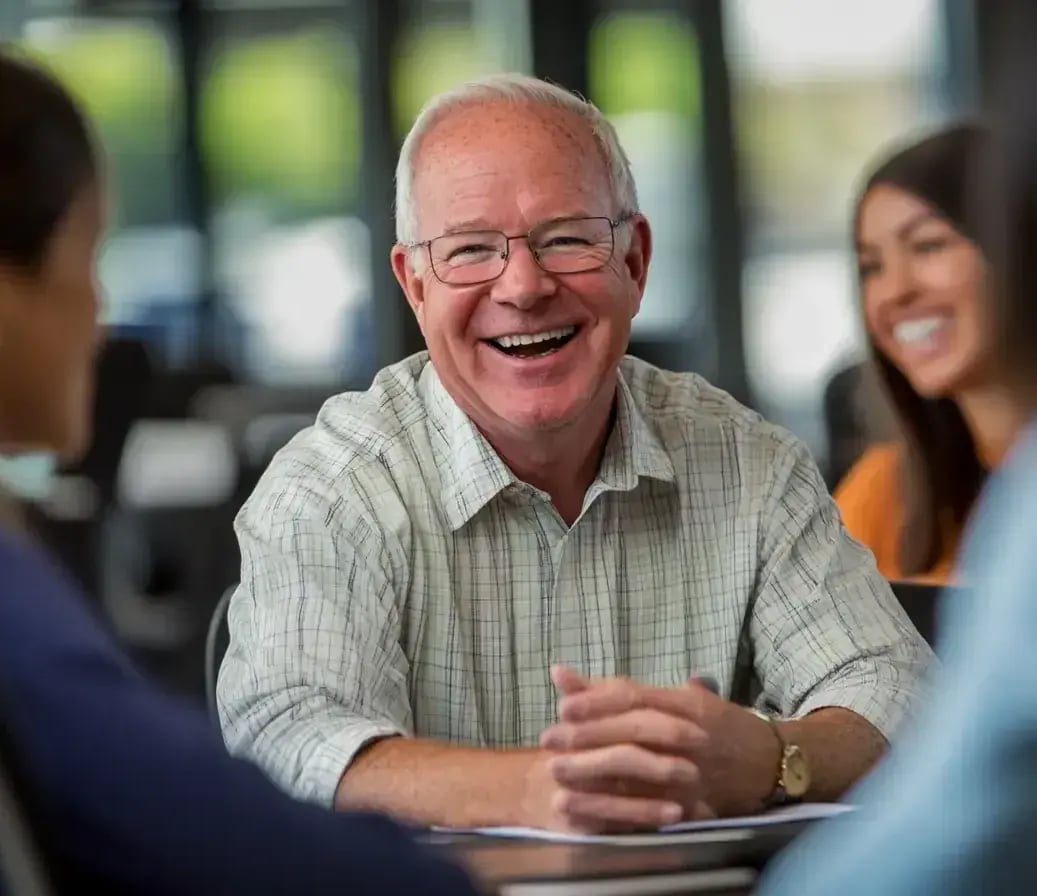 An older man in glasses and a plaid shirt laughs openly while chatting with colleagues during a group discussion. His relaxed posture and genuine smile add to the friendly energy in the room.